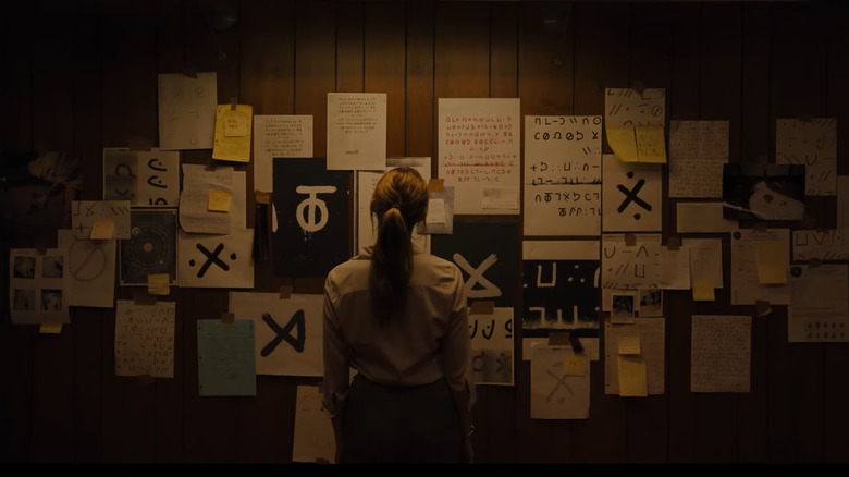 Un agent FBI care stă în fața unui perete, acoperit cu un colaj de litere, glife misterioase și alte accesorii ale unui caz de crimă din Longlegs