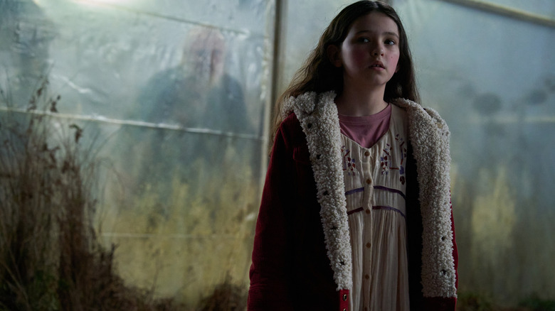 Matilda Firth în rolul lui Ginger stând într-o seră, în timp ce o figură ascunsă se strecoară în spatele ei în Wolf Man