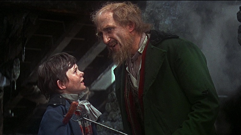 Fagin rânjește în timp ce ține un cârnați pe un băț în fața lui Dodger din Oliver!