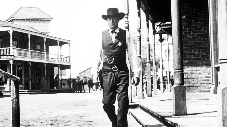 Gary Cooper în calitate de mareșal Will Kane, purtând o pălărie de cowboy neagră, o vestă și o cămașă și o stea a unui mareșal, ține un pistol în mâna dreaptă și se află în strada principală a unui oraș mic