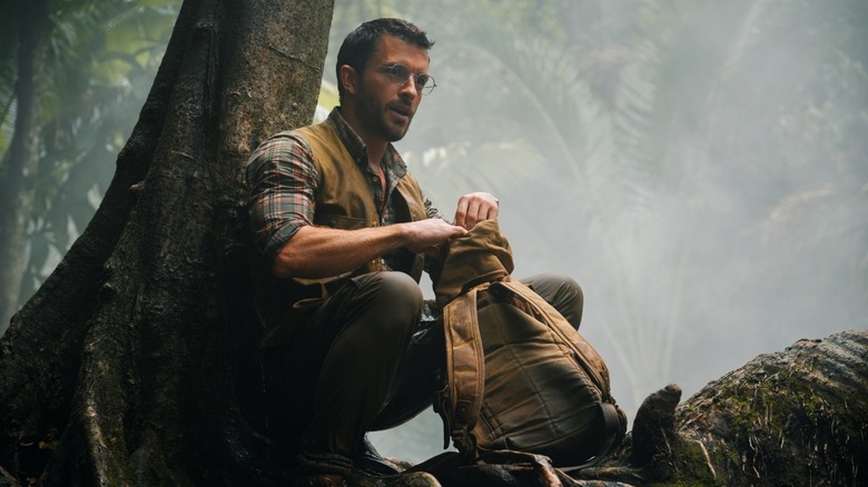Dr. Henry Loomis stând pe un copac cu rucsacul în fața lui pe pământ în Jurassic World Rebirth
