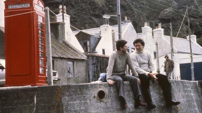 Peter Riegert ca Mac așezat pe un perete port lângă o cutie telefonică roșie în eroul local