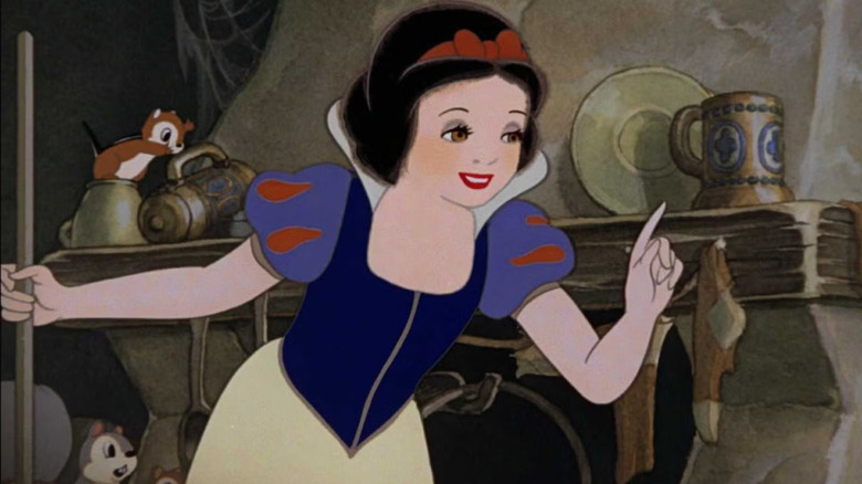 Albă ca zăpada ține o mătură și își ridică degetul indicatorului în timp ce vorbește în interiorul căsuței pitice prăfuite, înfundate în Albă ca Zăpada și cei șapte pitici