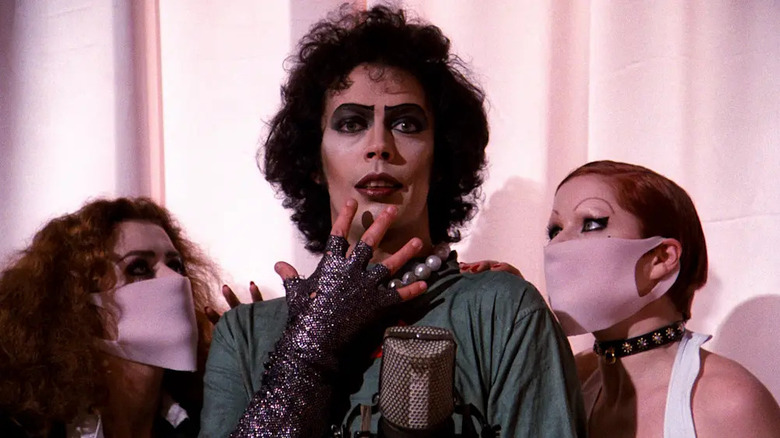 Frank-N-Furter într-o rochie de chirurgi stătea în fața unui microfon cu Columbia și Magenta de o parte