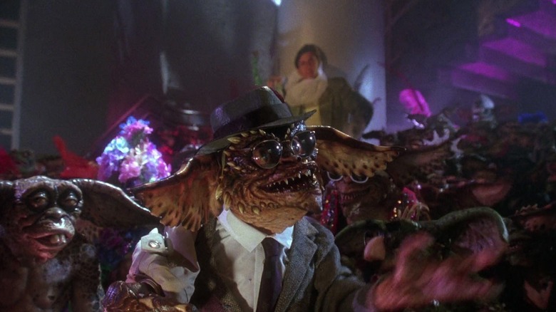 Brainy Gremlin, purtând o pălărie și ochelari în timp ce este îmbrăcat într -un costum formal și cravată, se adresează mulțimii care se petrece de gremlins în 