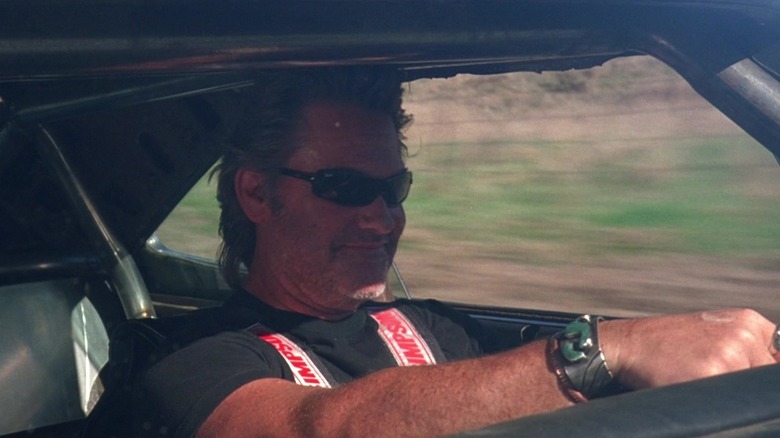 Kurt Russell ca cascador Mike, purtând ochelari de soare, conducând și zâmbind în dovada morții