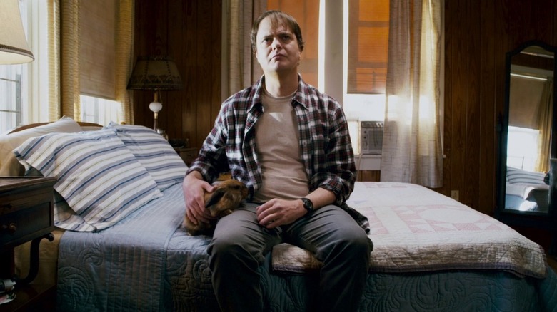 Rainn Wilson în rolul lui Frank Darbo în blugi și flanel, așezat pe patul său și făcând un iepure, în super