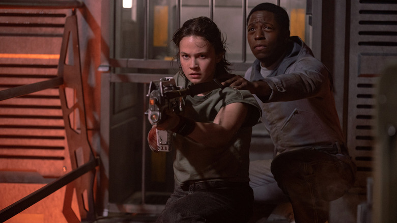 Cailee Spaeny ca ploaie ținând o armă futuristă, în timp ce David Jonsson, în timp ce Andy se ghemuiește protejat în spatele ei în Alien: Romulus