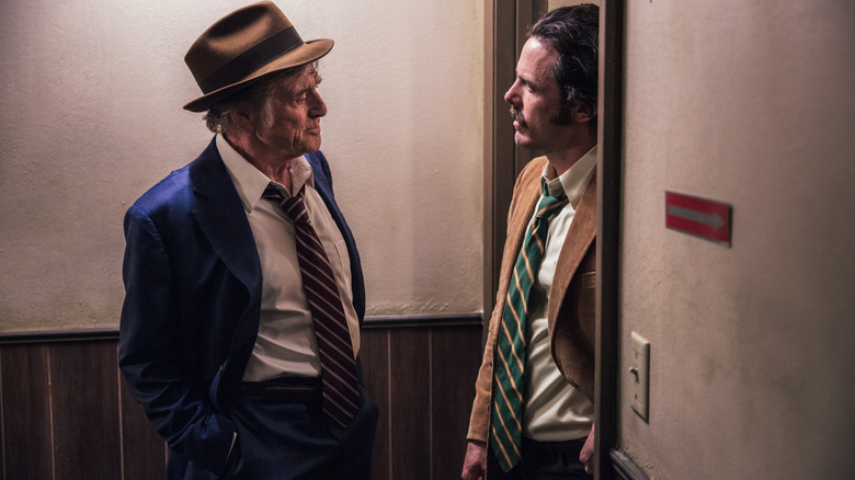 Robert Redford ca Forrest Tucker s -a îmbrăcat într -o pălărie de sus și un costum stând lângă o ușă de lângă Casey Affleck ca detectivul John Hunt în Old Man & the Gun