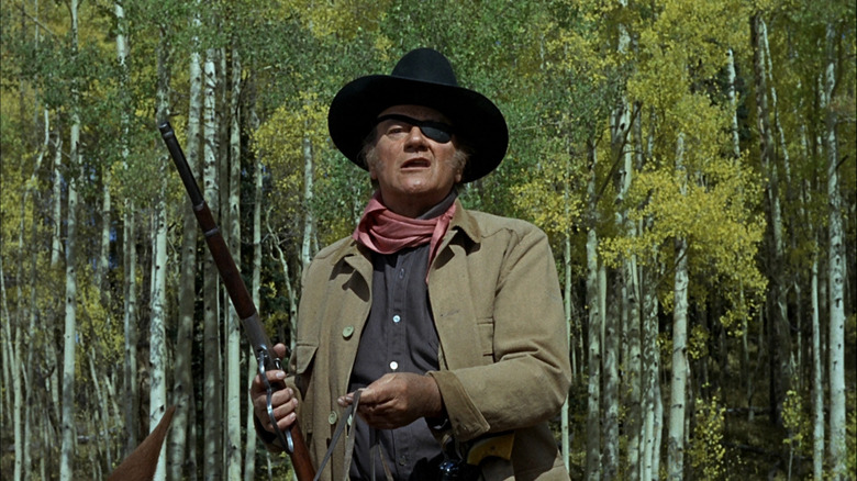 Cocoșul lui John Wayne Cogburn ține o armă în timp ce stă pe calul său în True Grit (1969)