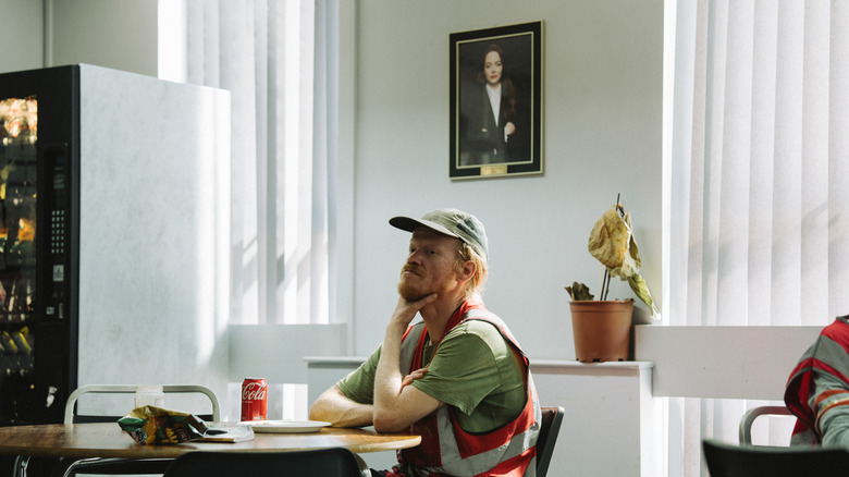 Teddy stă într -o sală de pauză sub o poză cu Michelle în Bugonia