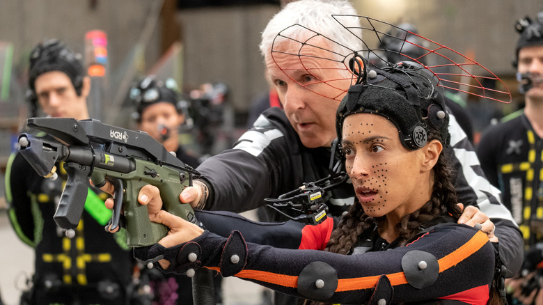 Regizorul James Cameron stând lângă Oona Chaplin în costumul ei de captare a mișcării în rolul lui Varang și ținând în mână o armă pe platoul Avatar: Fire & Ash