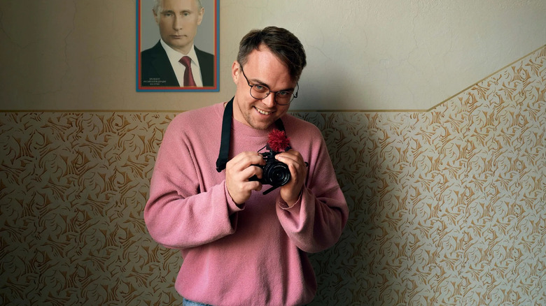 Pavel Talankin zâmbește răutăcios și ține o cameră în fața unei fotografii cu Vladimir Putin din Mr. Nobody Against Putin
