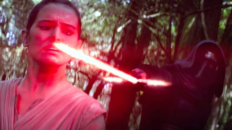Daisy Ridley în rolul lui Rey, cu Adam Driver în rolul lui Kylo Ren ținându-și sabia laser în față în Star Wars: The Force Awakens