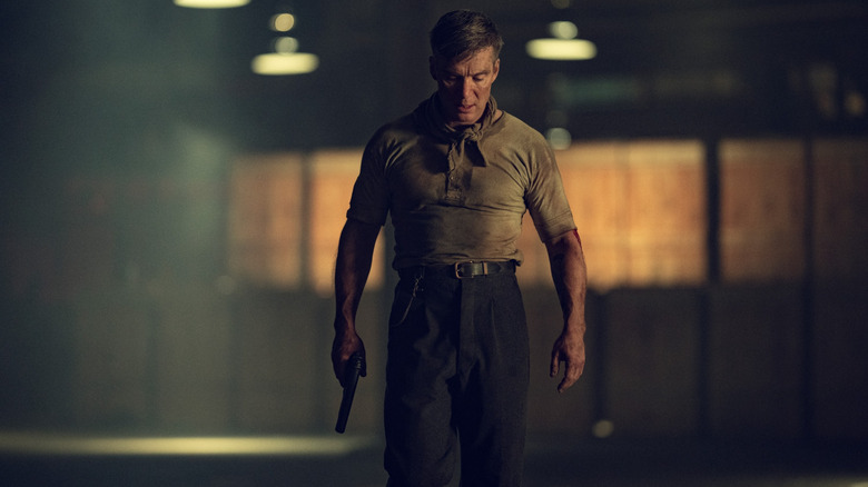 Cillian Murphy în rolul lui Tommy Shelby acoperit de pământ și ținând un pistol în mână în timp ce trece printr-un depozit gol în Peaky Blinders: The Immortal Man