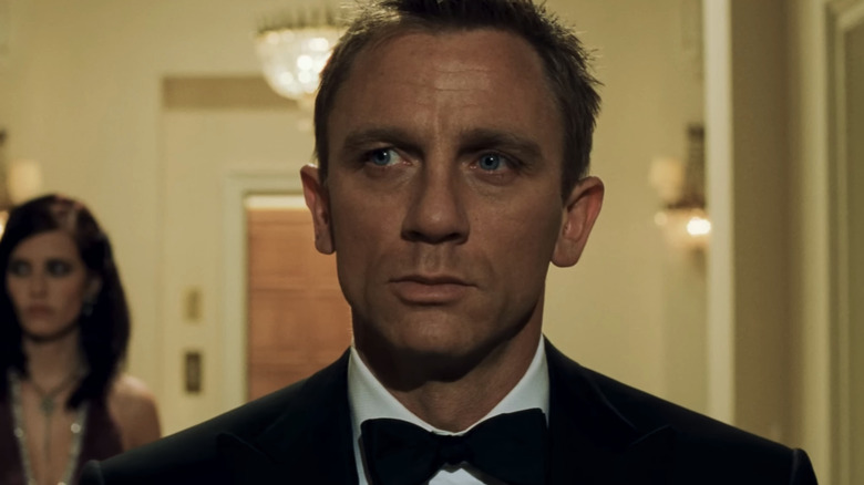 James Bond al lui Daniel Craig stă pe holul unui hotel, în timp ce Vesper Lynd, al lui Eva Green, stă în spatele lui în Casino Royale