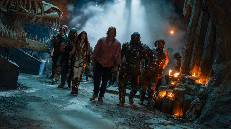 Nicholas Galitzine în rolul lui Adam care se plimbă printr-o cameră a unui castel plin de fum, alături de Camila Mendes în rolul Teela, Jon Xue Zhang în rolul Ram Man, Christiaan Bettridge în rolul lui Dian, Idris Elba în rolul Omului de arme și Johannes Haukur Johannesson în rolul lui Fisto în Masters of the Universe