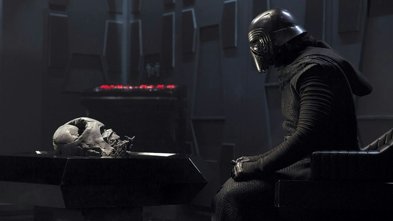 Adam Driver în rolul lui Kylo Ren stând pe un scaun vizavi de casca topită a lui Darth Vader în Star Wars: The Force Awakens