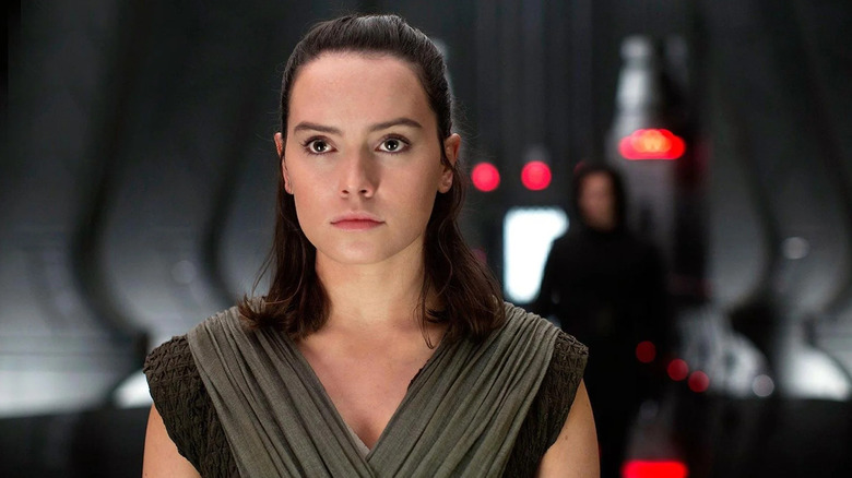 Daisy Ridley în rolul lui Rey stând într-o sală a tronului, cu Adam Driver în rolul lui Kylo Ren, lipsit de focalizare în spatele ei în Star Wars: The Last Jedi