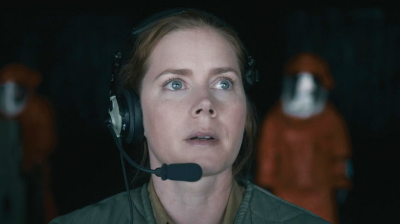 Louise Banks, de la Amy Adams, stă cu căștile puse în fața oamenilor îmbrăcați în costume cu materii periculoase la Sosire
