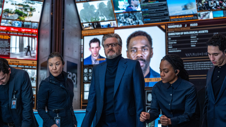 Colin Firth ca Noah Scanlon stând într-o cameră de control plină cu ecrane și alți angajați în Ziua Dezvăluirii