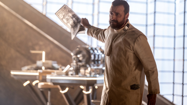 David Krumholtz în rolul lui Zor-El ținând o schemă și făcând semn spre banca lui de lucru în Supergirl