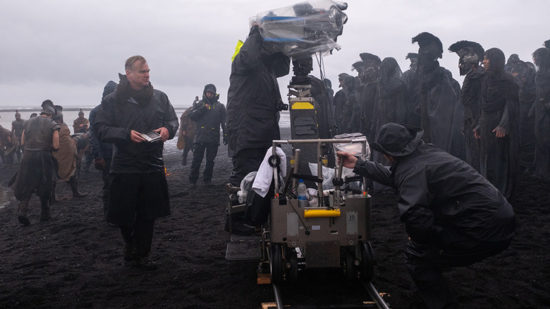 Regizorul Christopher Nolan purtând o jachetă de ploaie și stând lângă o cameră pe o plajă în fața mai multor figuranți de pe platoul Odiseei