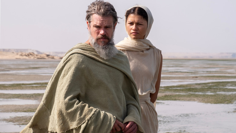 Matt Damon în rolul lui Ulise stând pe o câmpie vastă, cu Zendaya în rolul lui Athena în spatele lui în Odiseea