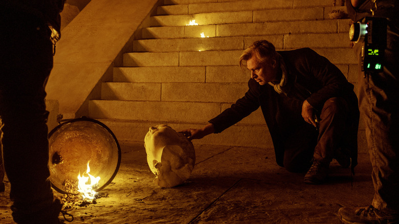 Regizorul Christopher Nolan ajustând cu atenție capul unei statui căzute în fața unei scări în platoul Odiseei