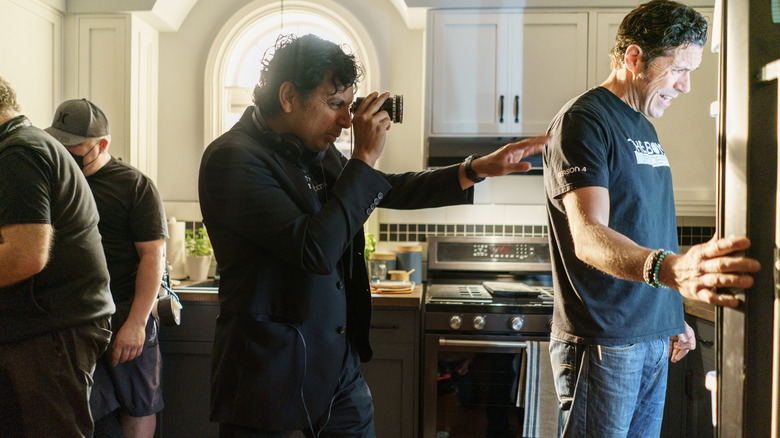 Regizorul M. Night Shyamalan stând într-o bucătărie alături de câțiva membri ai echipajului care desenează o fotografie pe platoul Trap