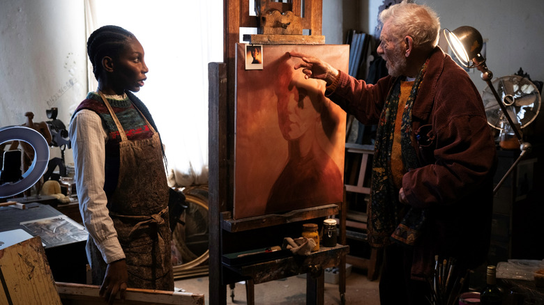 Michaela Coel în rolul lui Lori Butler, stând într-o cameră aglomerată lângă Ian McKellen, în rolul lui Julian Sklar, făcând semne către un tablou din The Christophers