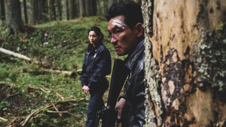Hwang Jung-min ca Bum-seok stând într-o pădure cu un bandaj pe cap lângă Hoyeon ca Sung-ae în Hope