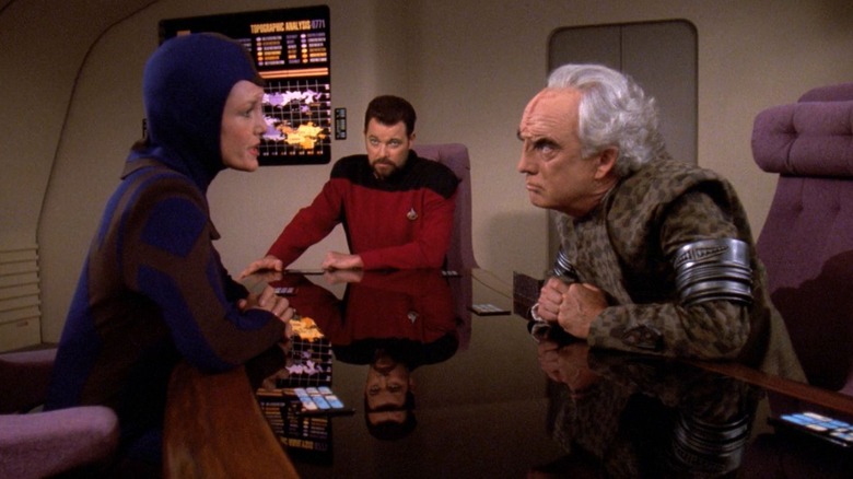 Lorin și Mauric se uită unul la altul peste o masă, cu Riker privind Star Trek: The Next Generation